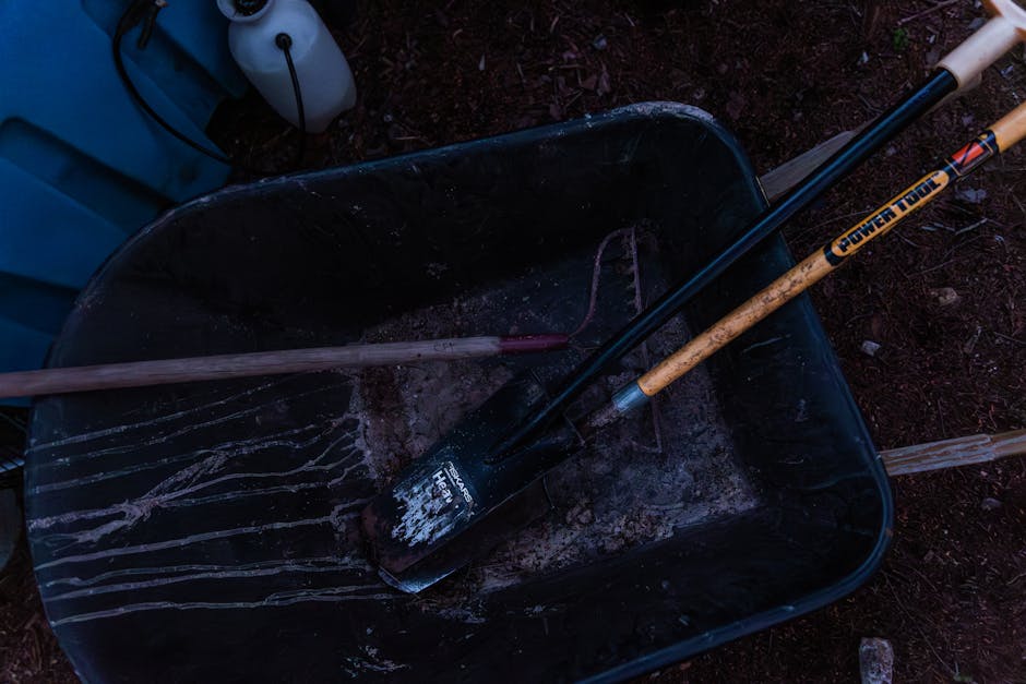 A dark shot of gardening tools including a shovel and rake in a wheelbarrow outdoors.