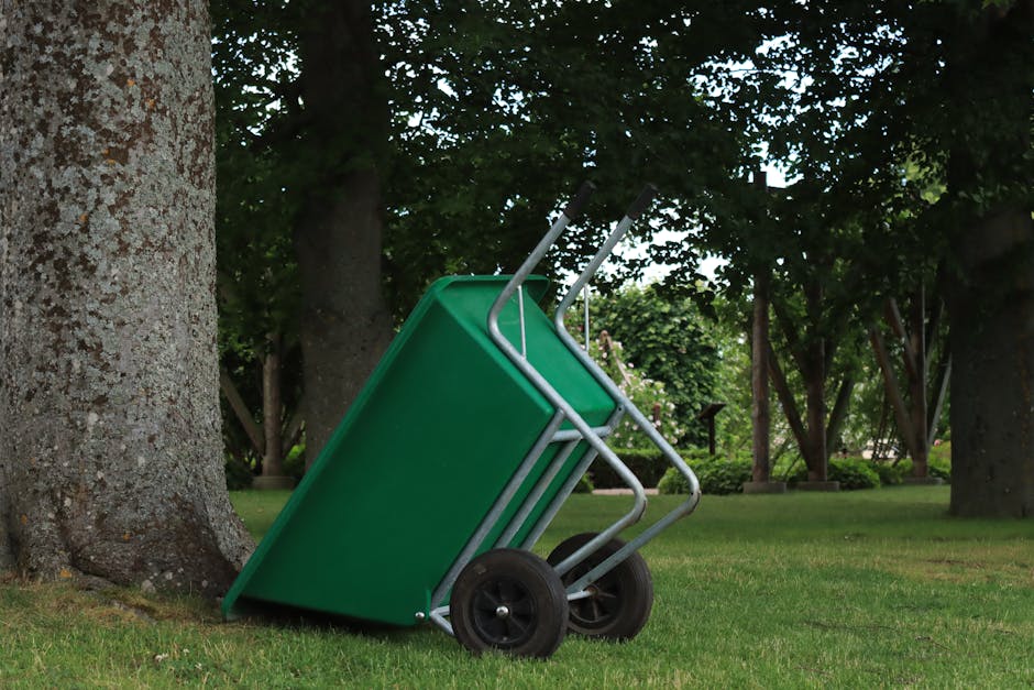 A green wheelbarrow leaning against a tree on a lush grassy lawn.